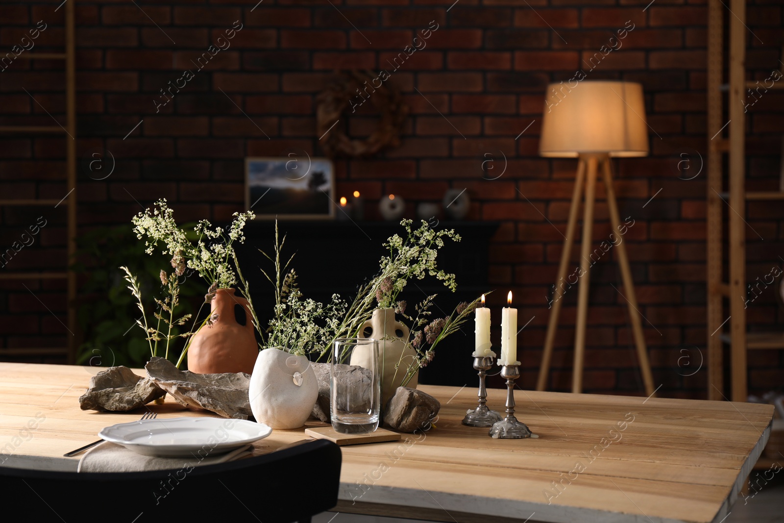 Photo of Set of clean dishware, vases with beautiful flowers, stones and burning candles on wooden table in stylish dining room