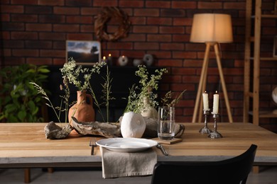 Photo of Set of clean dishware, vases with beautiful flowers, stones and burning candles on wooden table in stylish dining room