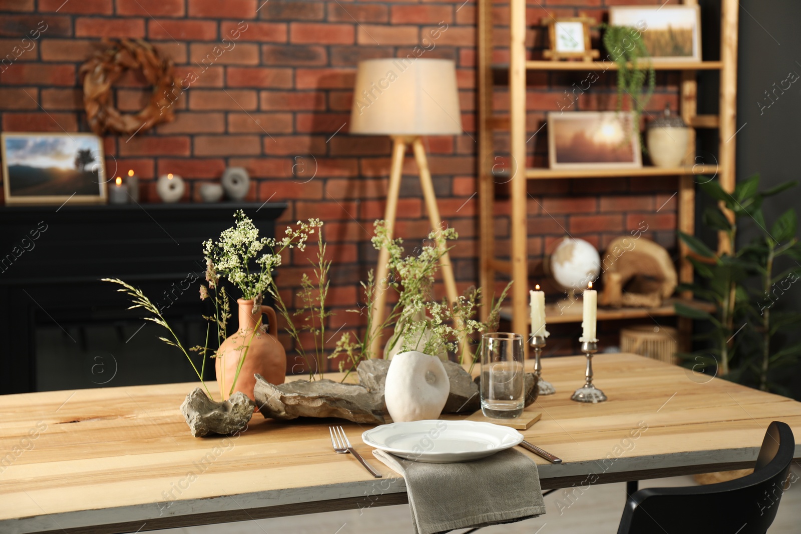 Photo of Set of clean dishware, vases with beautiful flowers, stones and burning candles on wooden table in stylish dining room