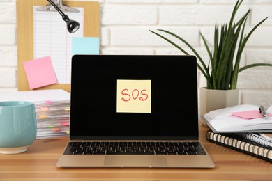 Photo of Note with word SOS, laptop and stationery on wooden table at workplace