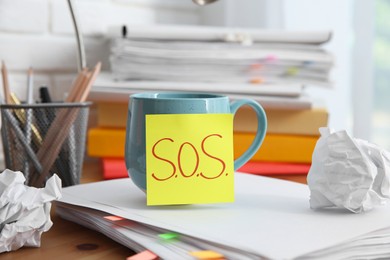 Photo of Note with word SOS, cup and stationery on wooden table at workplace