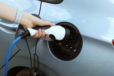 Photo of Woman inserting plug into electric car socket at charging station outdoors, closeup