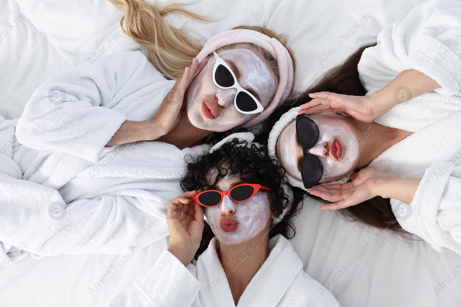 Photo of Happy friends with facial masks and sunglasses on bed, top view. Spa party