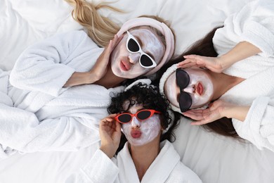 Photo of Happy friends with facial masks and sunglasses on bed, top view. Spa party