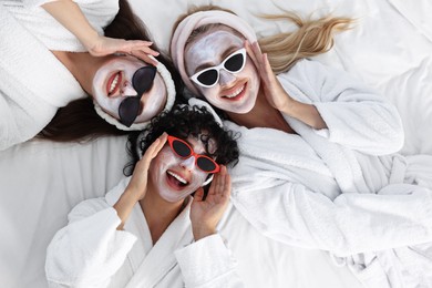 Photo of Happy friends with facial masks and sunglasses on bed, top view. Spa party