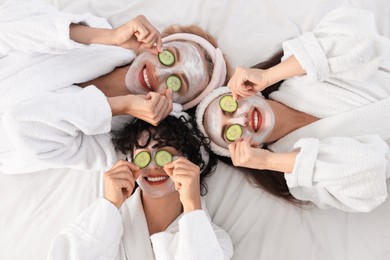 Photo of Happy friends with facial masks and cucumber slices on bed, top view. Spa party