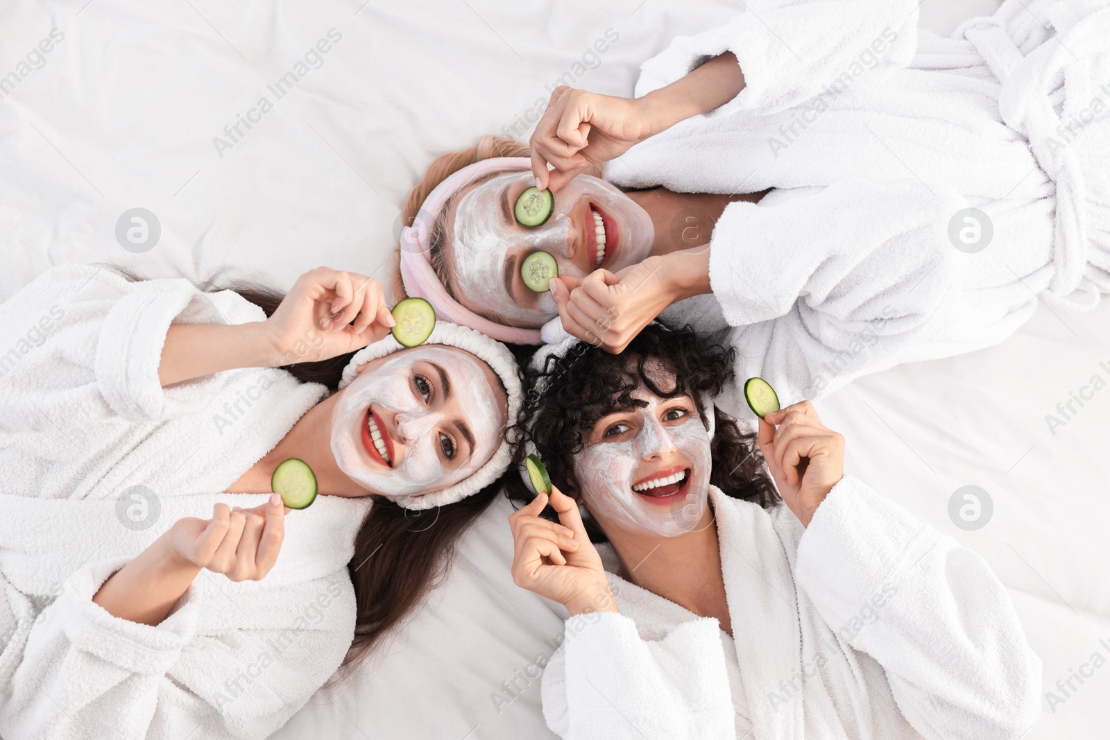 Photo of Happy friends with facial masks and cucumber slices on bed, top view. Spa party