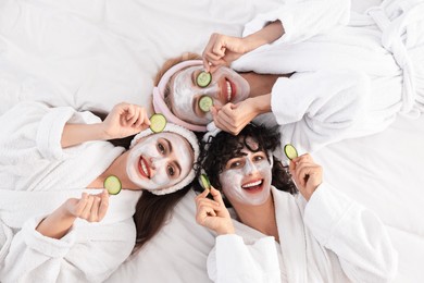Photo of Happy friends with facial masks and cucumber slices on bed, top view. Spa party