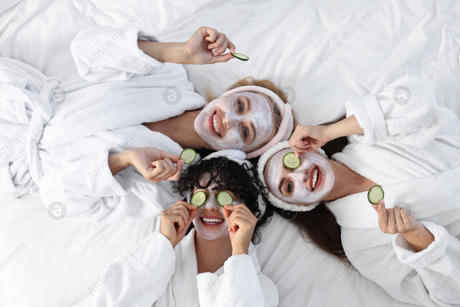 Photo of Happy friends with facial masks and cucumber slices on bed, top view. Spa party