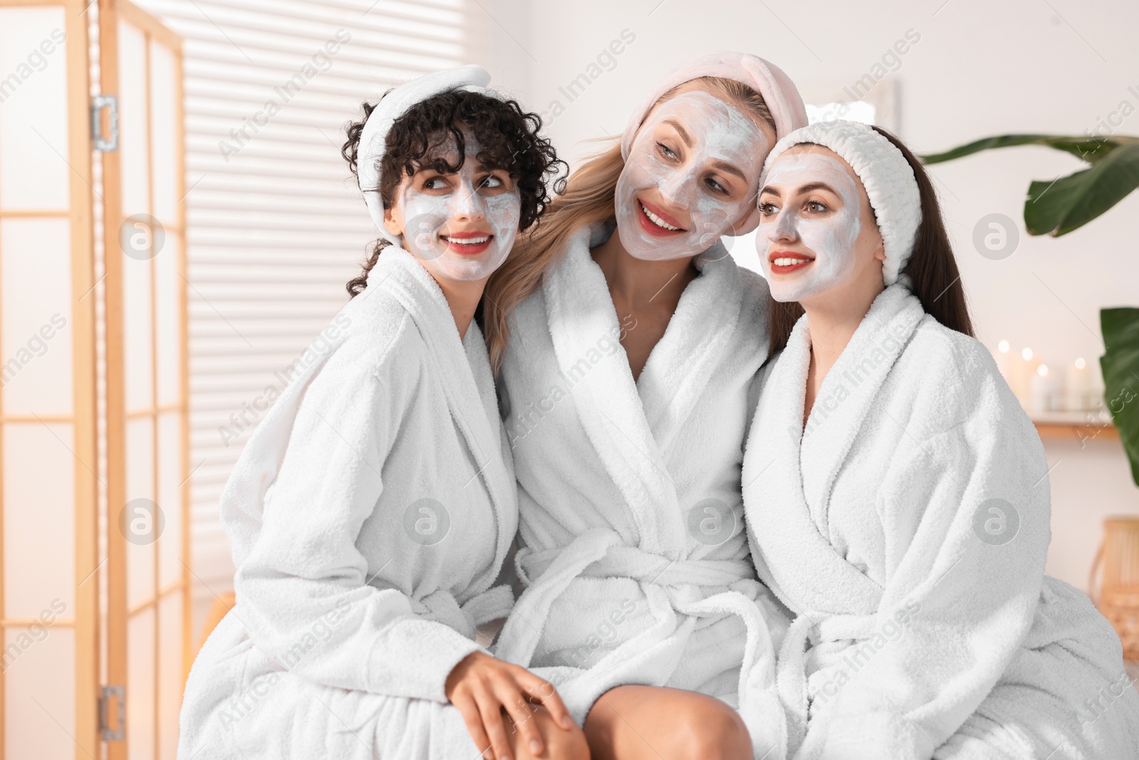 Photo of Happy friends in bathrobes with facial masks indoors. Spa party