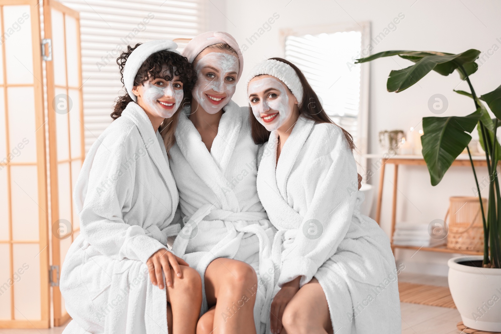 Photo of Happy friends in bathrobes with facial masks indoors. Spa party