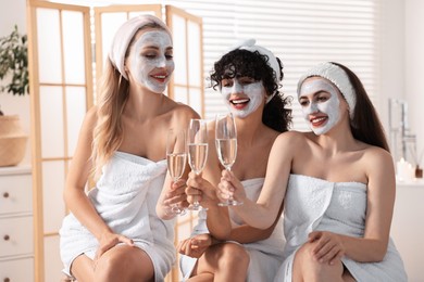 Photo of Happy friends with facial masks and glasses of sparkling wine in bathroom. Spa party