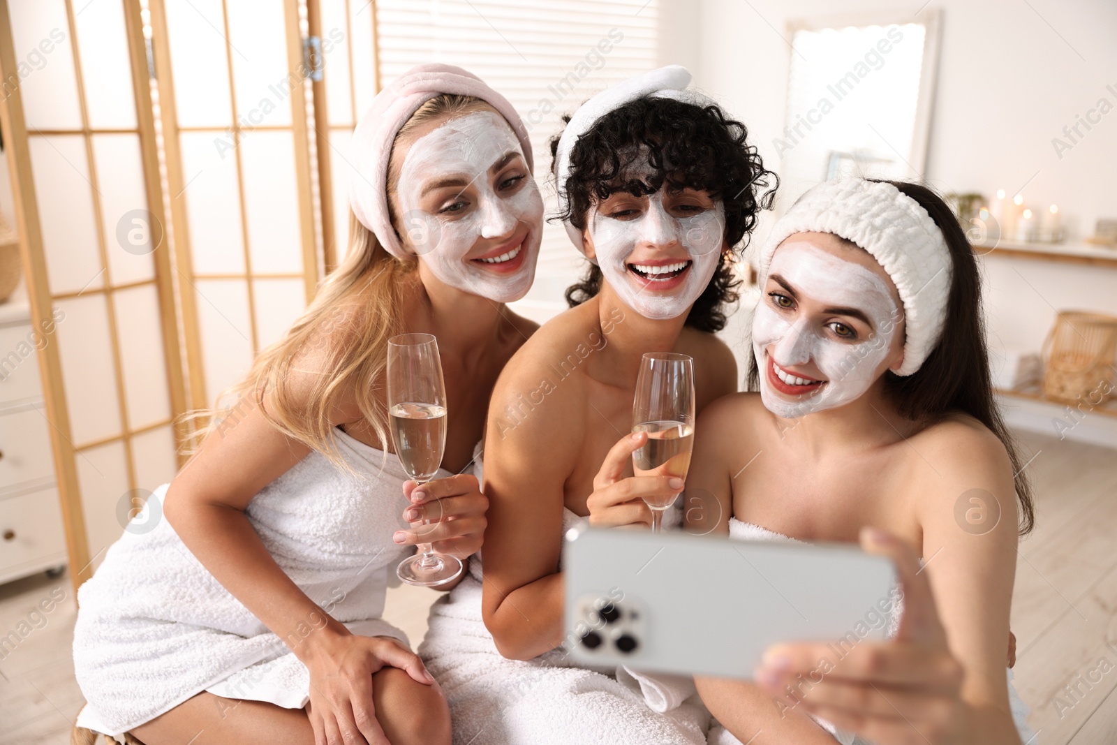 Photo of Happy friends with facial masks and glasses of sparkling wine taking selfie in bathroom. Spa party