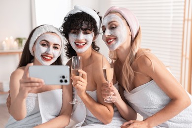 Photo of Happy friends with facial masks and glasses of sparkling wine taking selfie in bathroom. Spa party
