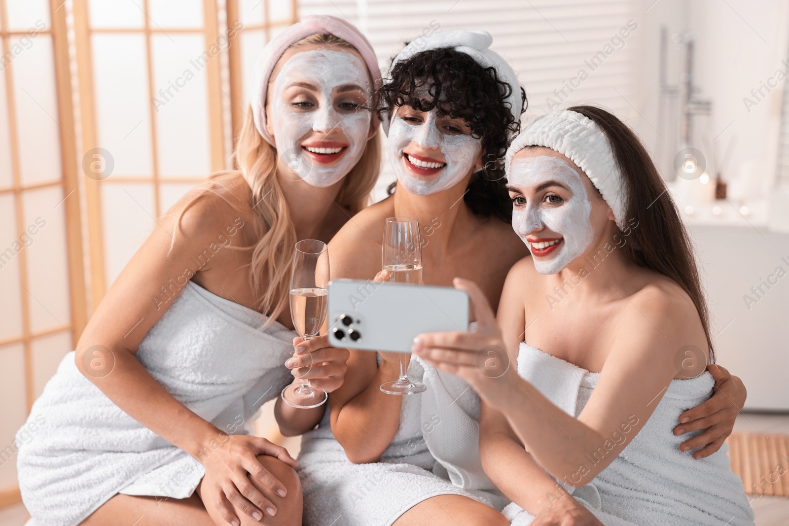 Photo of Happy friends with facial masks and glasses of sparkling wine taking selfie in bathroom. Spa party