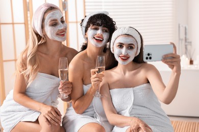 Happy friends with facial masks and glasses of sparkling wine taking selfie in bathroom. Spa party