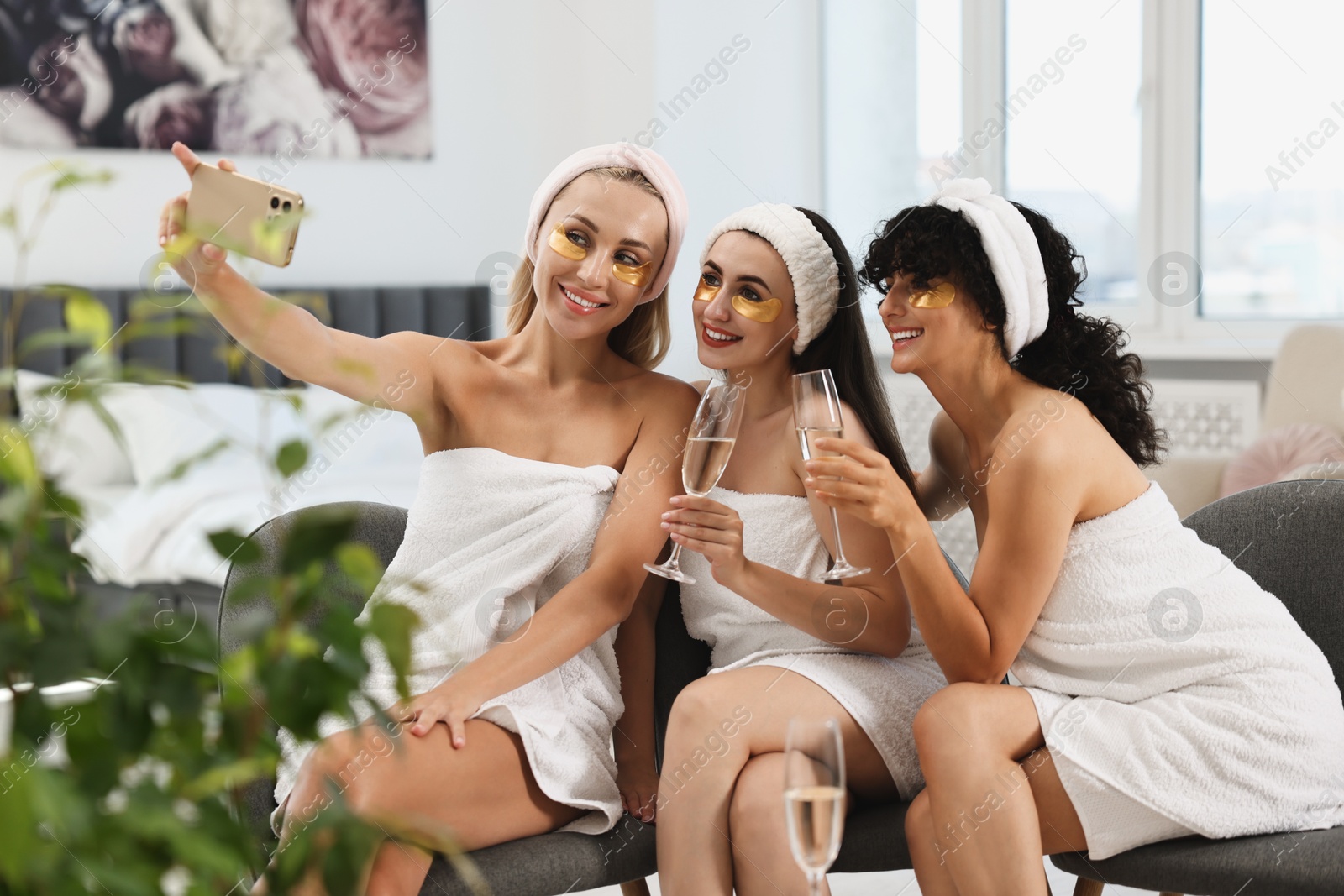 Photo of Happy friends with glasses of sparkling wine taking selfie indoors. Spa party