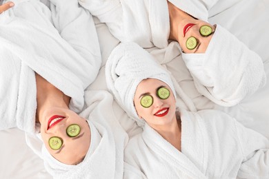 Happy friends in bathrobes with cucumber slices on bed, top view. Spa party