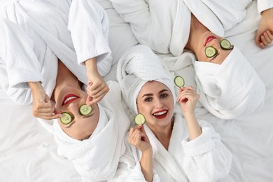 Happy friends in bathrobes with cucumber slices on bed, top view. Spa party