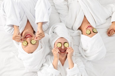 Happy friends in bathrobes with cucumber slices on bed, top view. Spa party