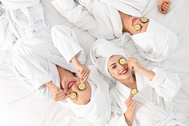 Happy friends in bathrobes with cucumber slices on bed, top view. Spa party