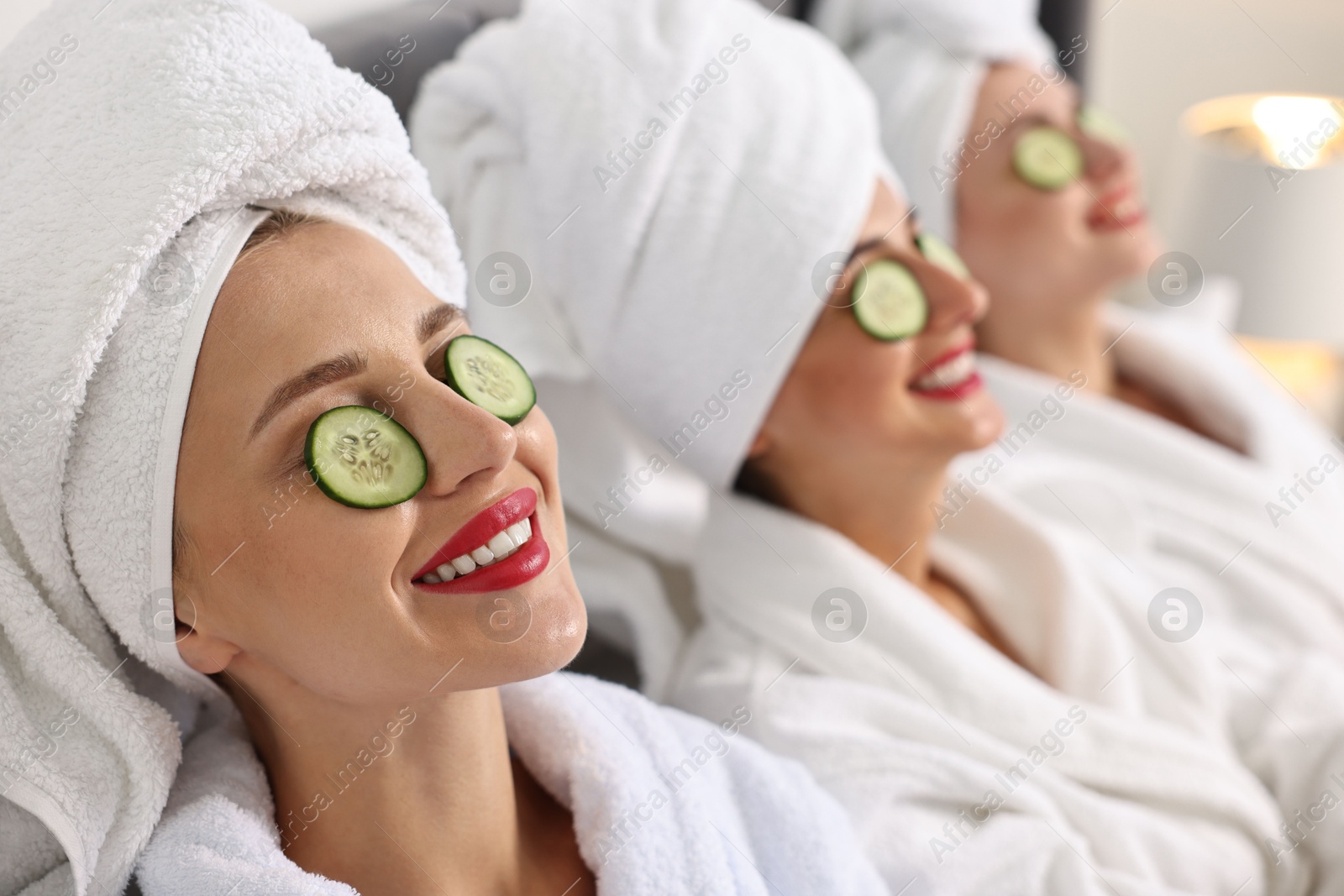 Photo of Happy friends in bathrobes with cucumber slices on bed, selective focus. Spa party