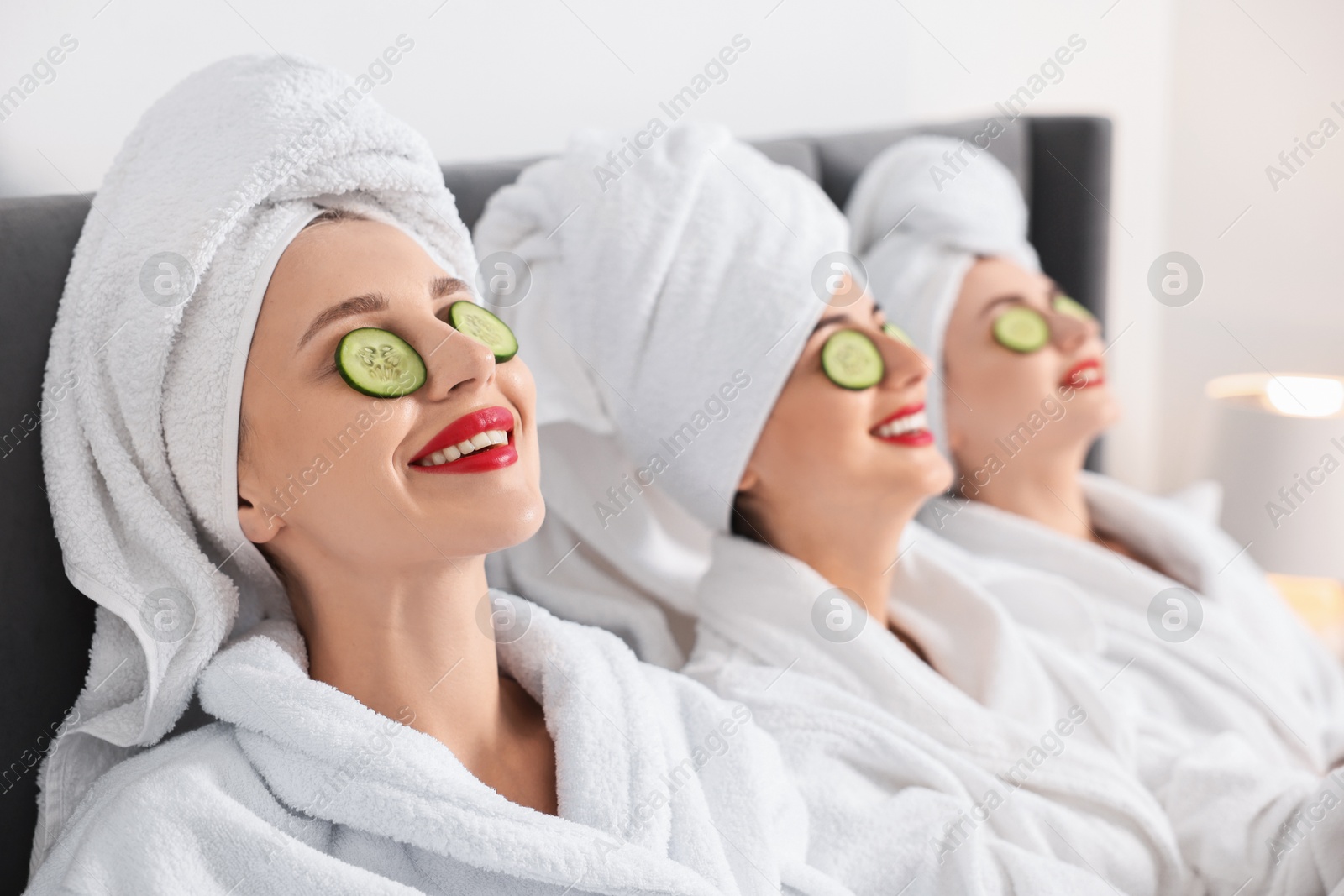 Photo of Happy friends in bathrobes with cucumber slices on bed, selective focus. Spa party