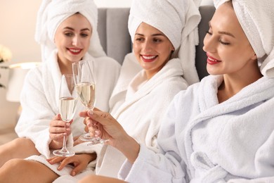 Photo of Happy friends in bathrobes with glasses of sparkling wine on bed, selective focus. Spa party
