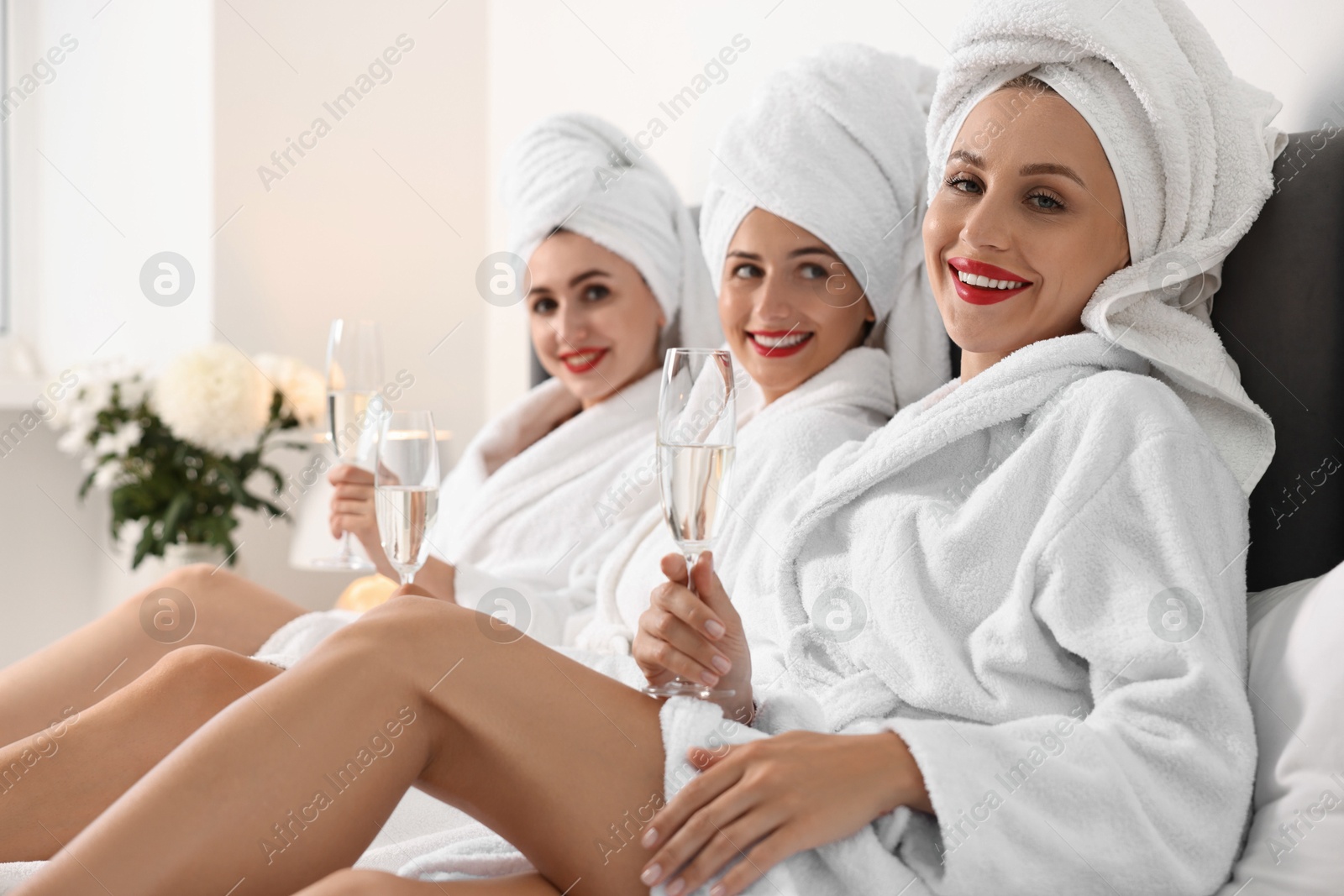 Photo of Happy friends in bathrobes with glasses of sparkling wine on bed, selective focus. Spa party