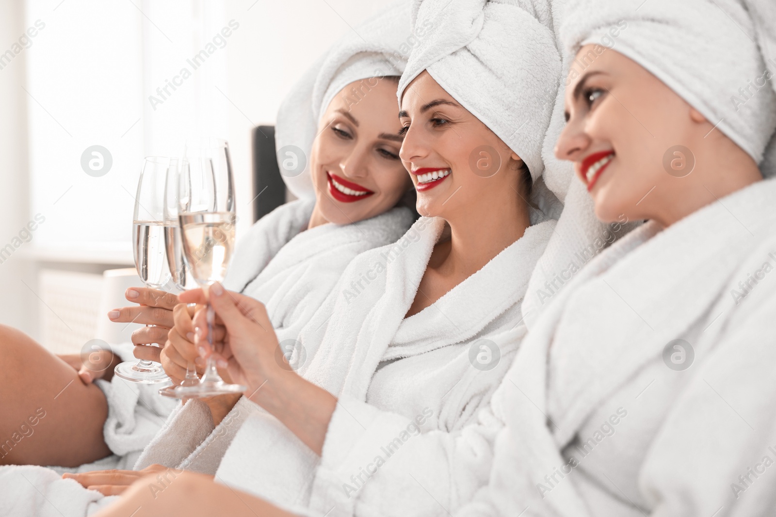 Photo of Happy friends in bathrobes with glasses of sparkling wine on bed, selective focus. Spa party