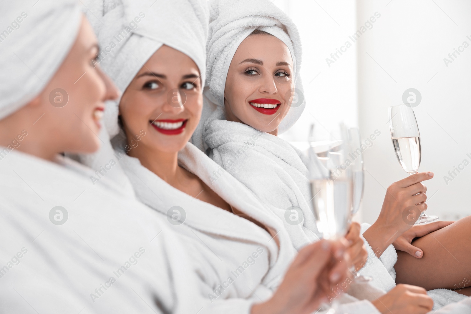 Photo of Happy friends in bathrobes with glasses of sparkling wine on bed, selective focus. Spa party