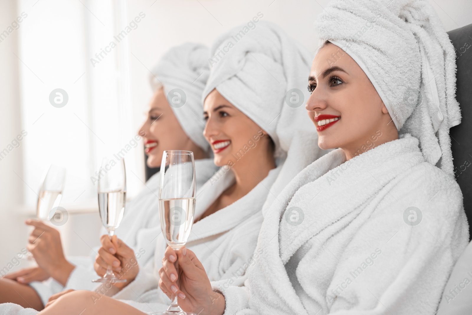 Photo of Happy friends in bathrobes with glasses of sparkling wine on bed, selective focus. Spa party