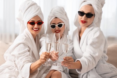 Photo of Happy friends in bathrobes with sunglasses and glasses of sparkling wine indoors, selective focus. Spa party