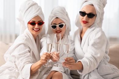 Happy friends in bathrobes with sunglasses and glasses of sparkling wine indoors, selective focus. Spa party