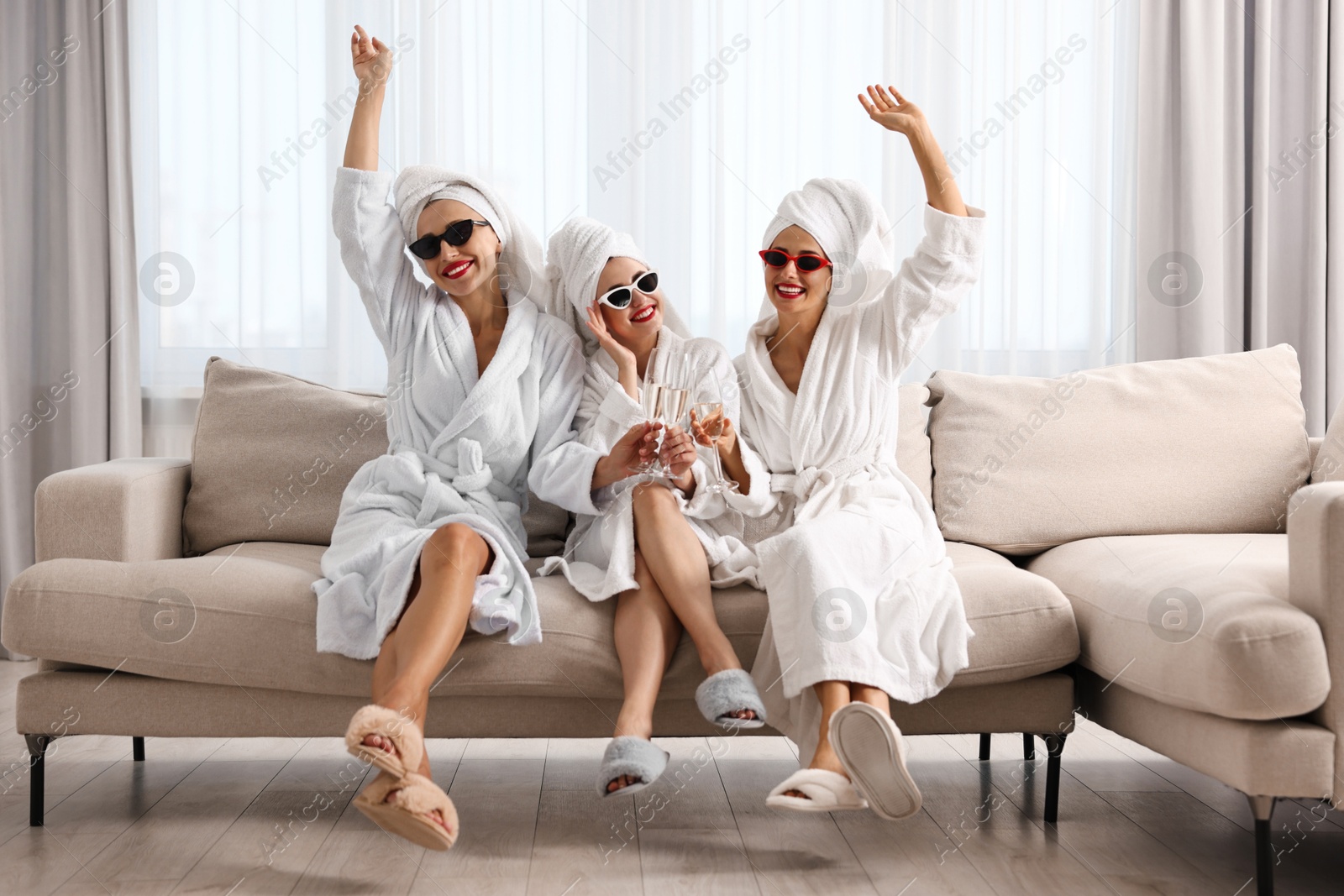 Photo of Happy friends in bathrobes with sunglasses and glasses of sparkling wine indoors. Spa party