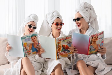 Photo of Happy friends in bathrobes with magazines on couch indoors. Spa party
