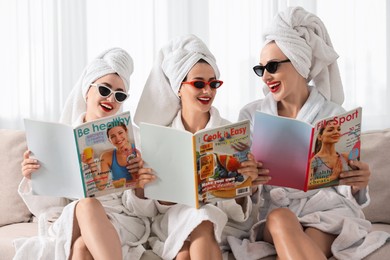 Happy friends in bathrobes with magazines on couch indoors. Spa party