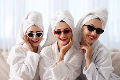 Happy friends in bathrobes with sunglasses on couch indoors. Spa party