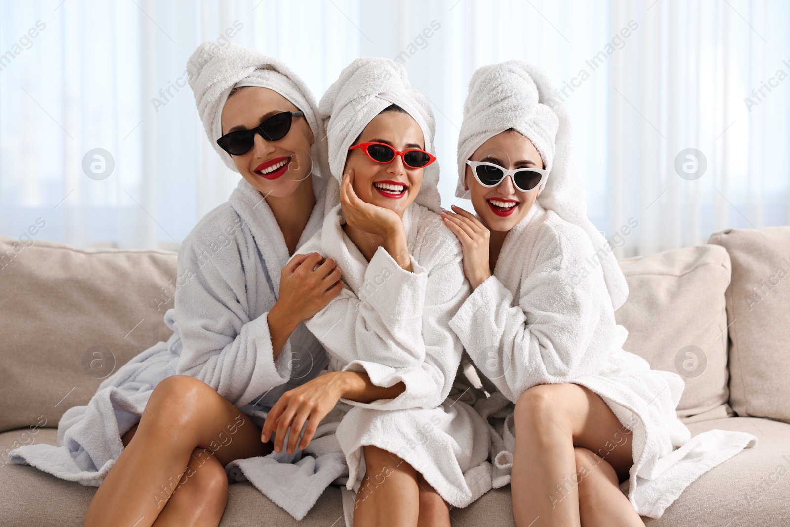 Photo of Happy friends in bathrobes with sunglasses on couch indoors. Spa party