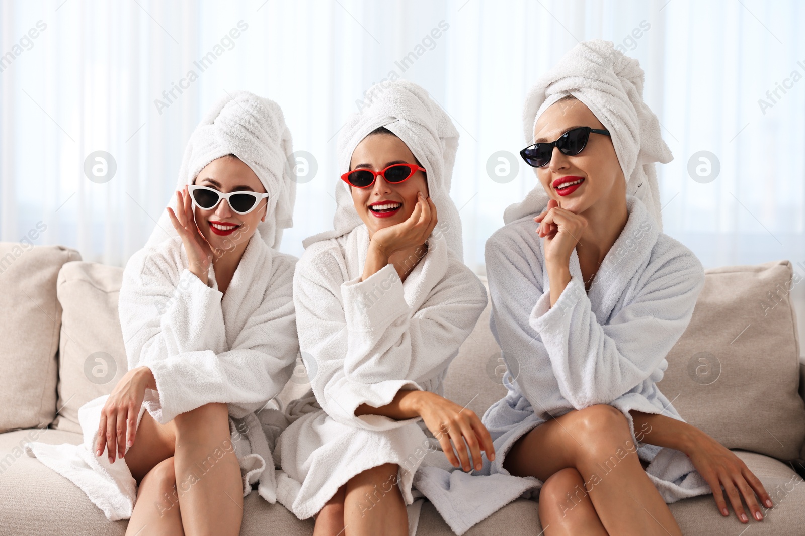 Photo of Happy friends in bathrobes with sunglasses on couch indoors. Spa party