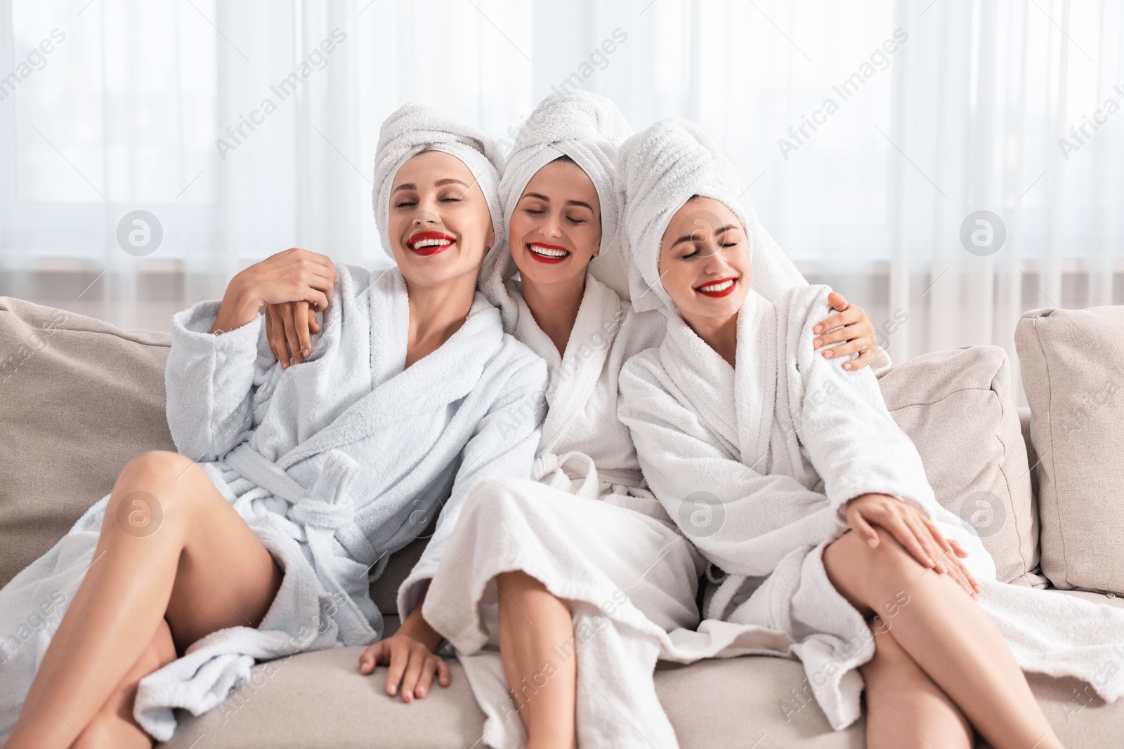 Photo of Happy friends in bathrobes with towels on couch indoors. Spa party