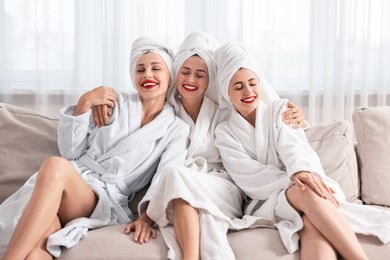 Photo of Happy friends in bathrobes with towels on couch indoors. Spa party
