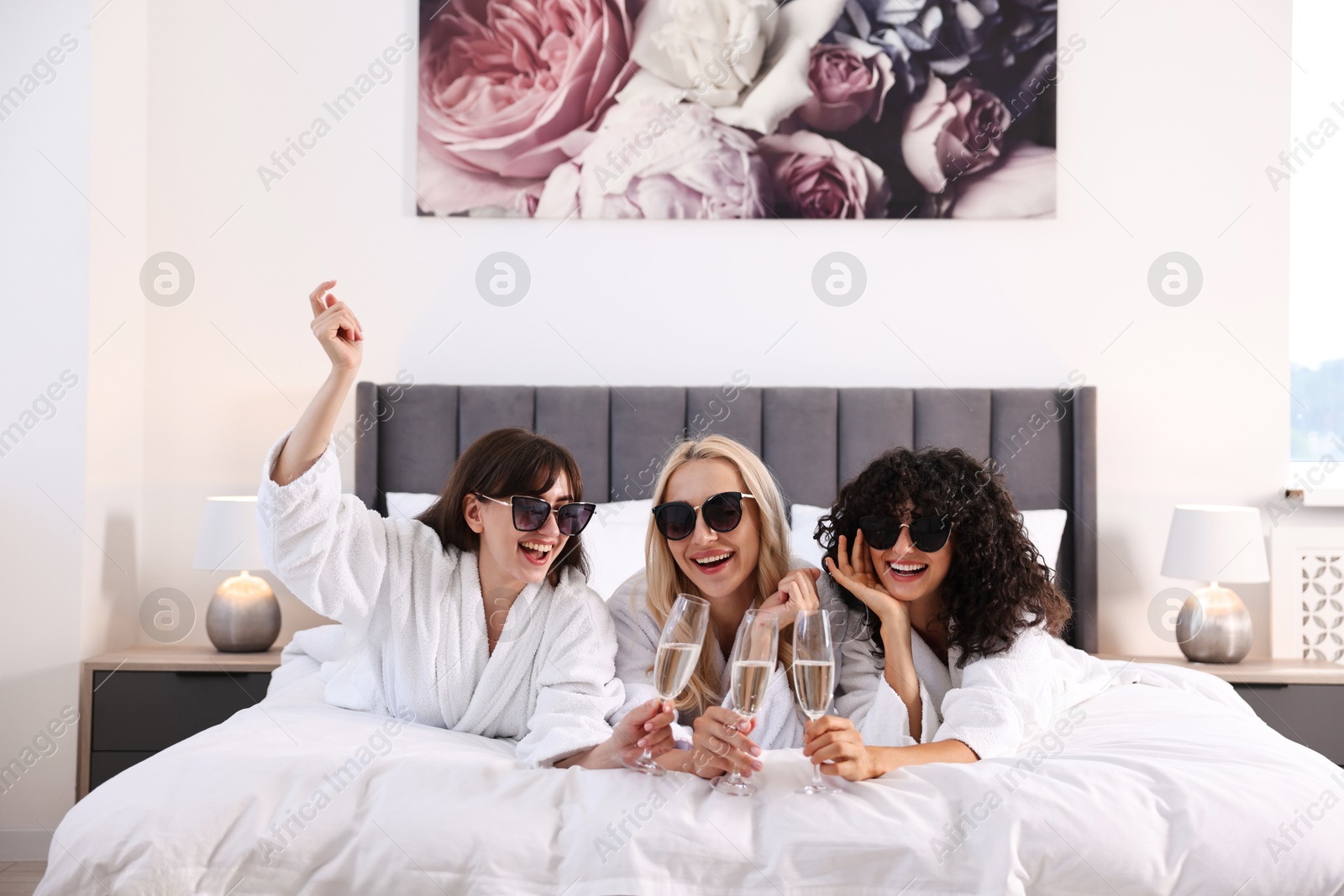 Photo of Happy friends with sunglasses and glasses of sparkling wine on bed. Spa party