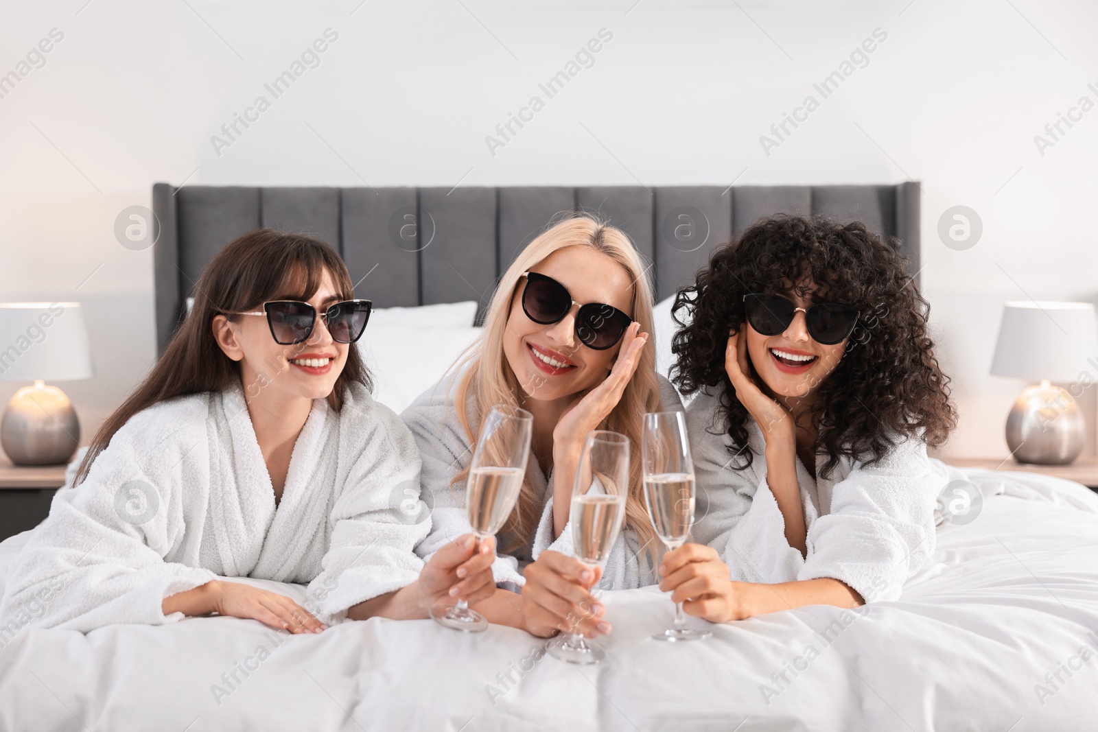 Photo of Happy friends with sunglasses and glasses of sparkling wine on bed. Spa party