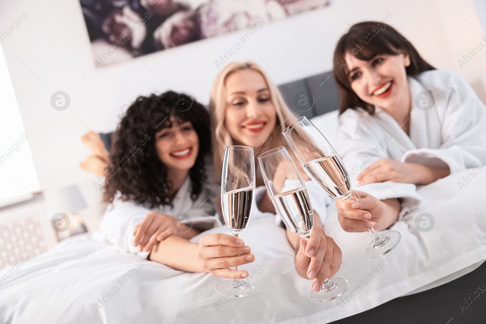 Photo of Happy friends clinking glasses of sparkling wine on bed, selective focus. Spa party