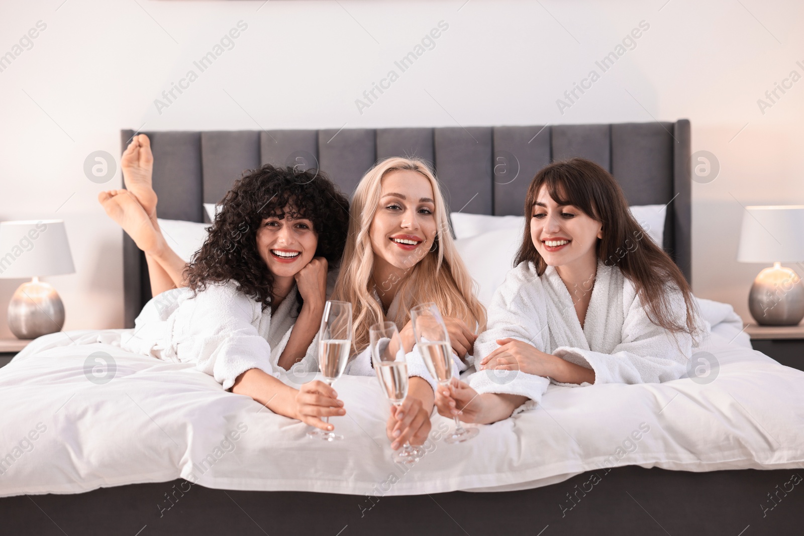 Photo of Happy friends with glasses of sparkling wine on bed. Spa party