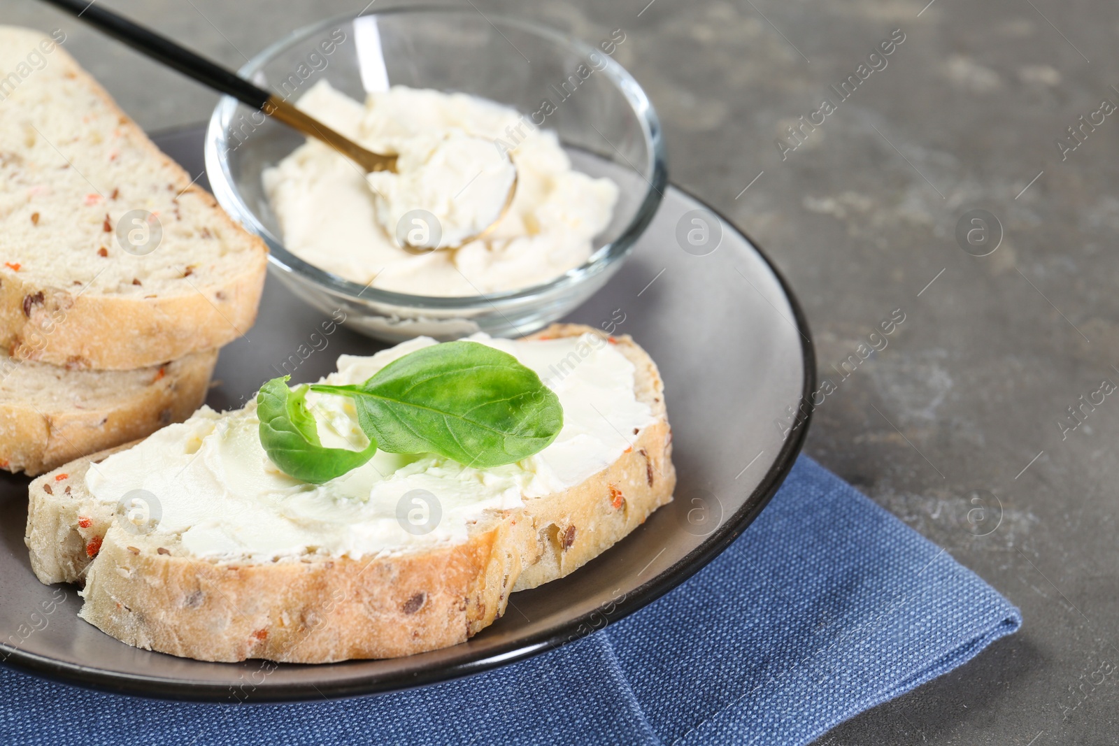 Photo of Pieces of bread with cream cheese and basil leaves on gray textured table, closeup