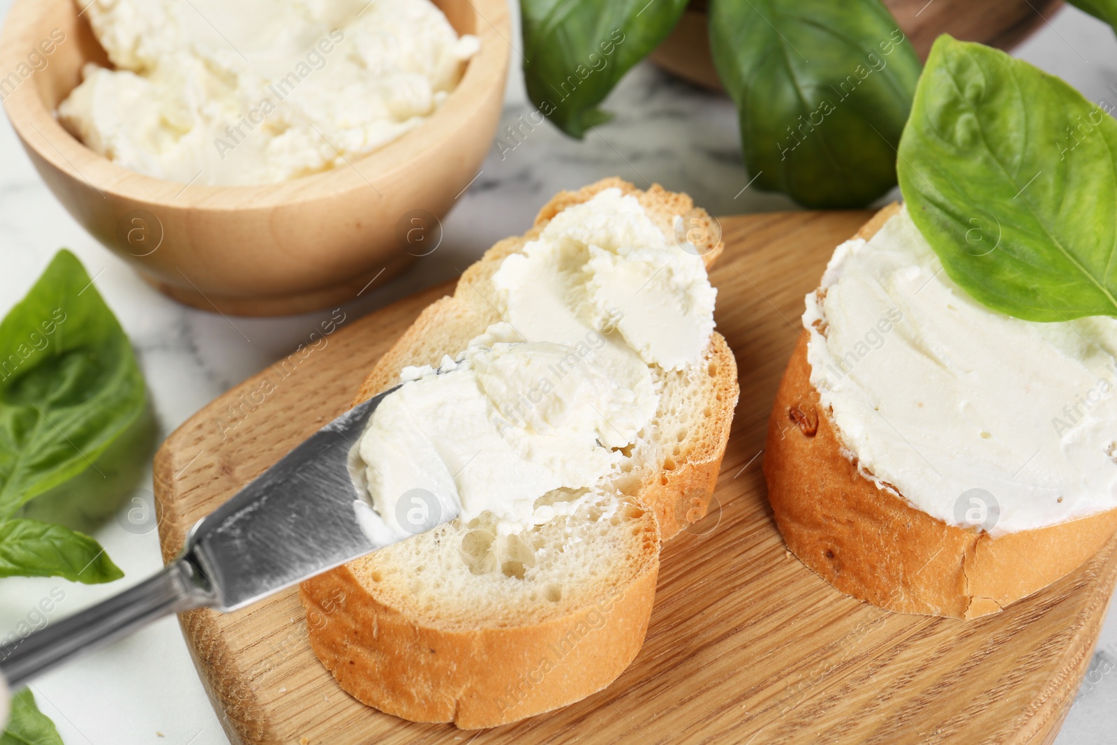 Photo of Pieces of bread with cream cheese, basil and knife on white table