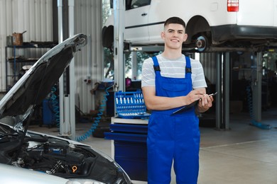 Young auto mechanic with clipboard taking notes near broken car at automobile repair shop