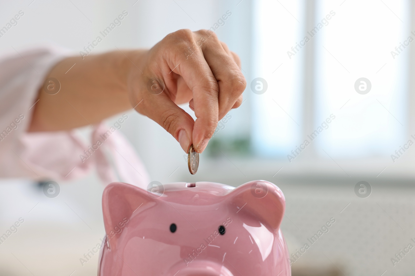 Photo of Woman putting coin into pink piggy bank at home, closeup